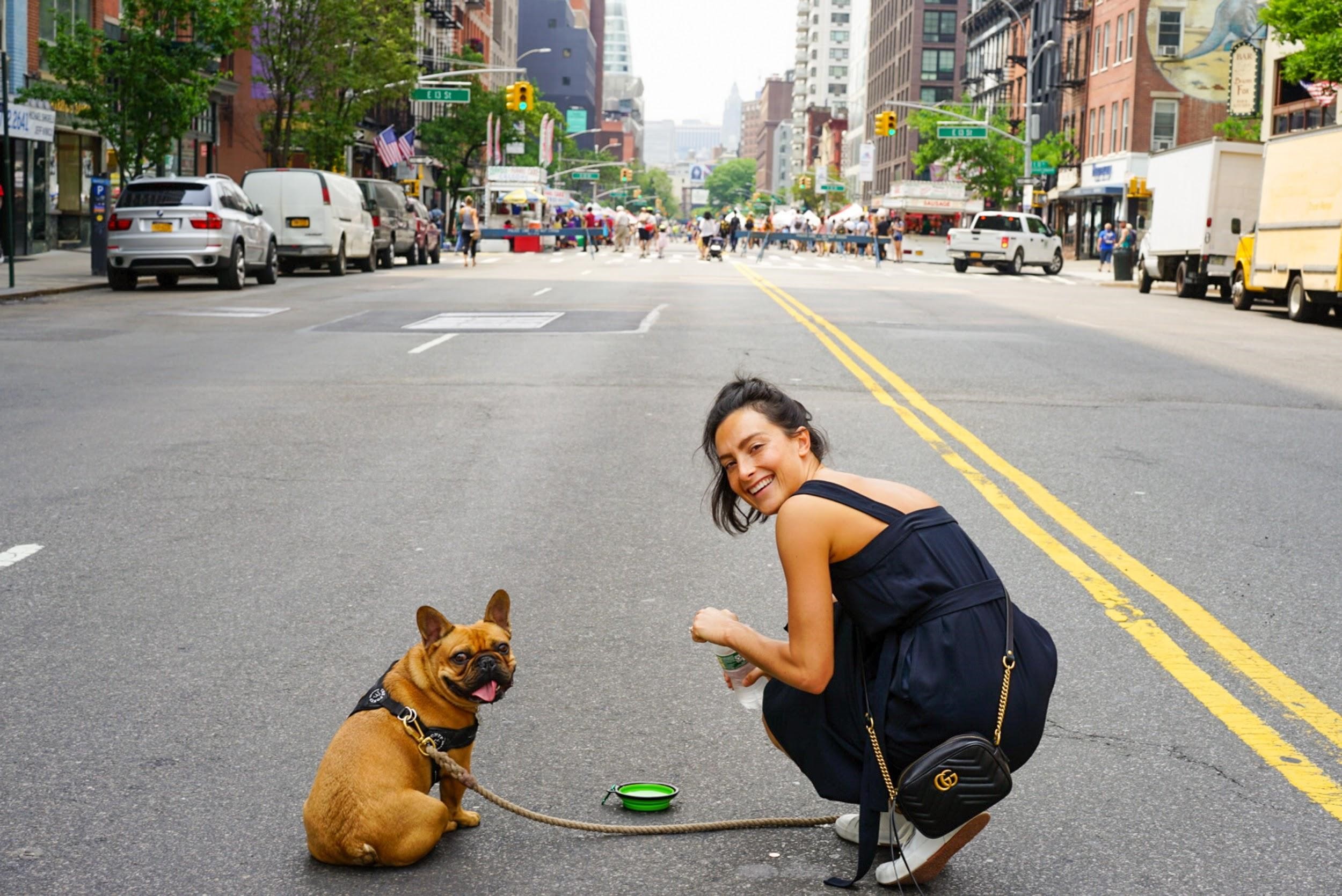 A girl and her French Bulldog is posing on the road in a large city.